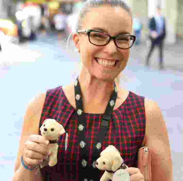 A person smiling at the camera while holding two mini Guide Dog plush puppies.